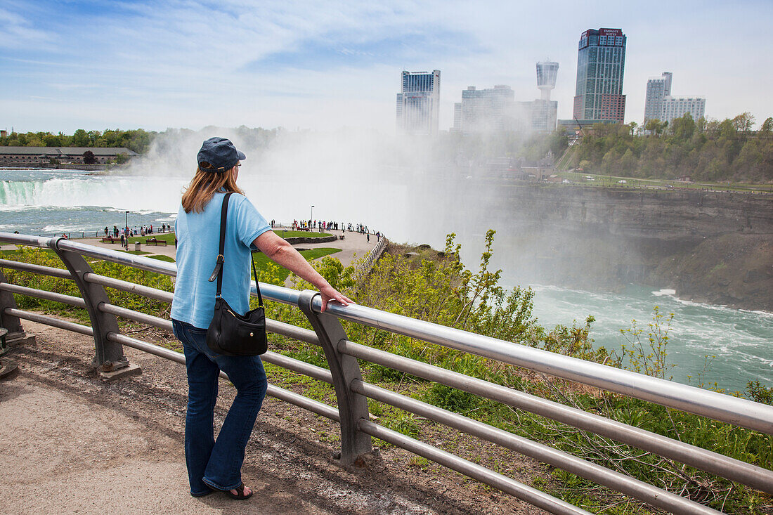 Eine Frau steht an einem Geländer mit Blick auf die Horseshoe Falls im Niagara Falls State Park im Bundesstaat New York, mit der Stadt Niagara Falls, Ontario im Hintergrund; Niagara Falls, New York, Vereinigte Staaten von Amerika