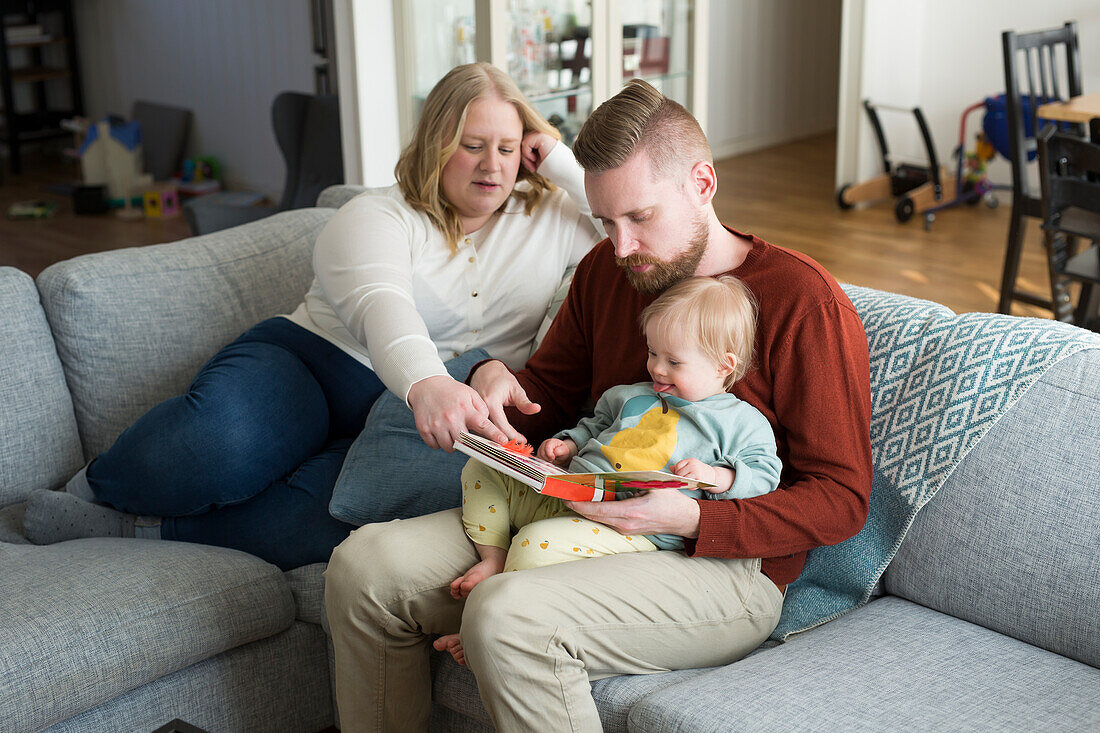 Eltern mit Baby mit Down-Syndrom lesen ein Buch auf Sofa im Wohnzimmer