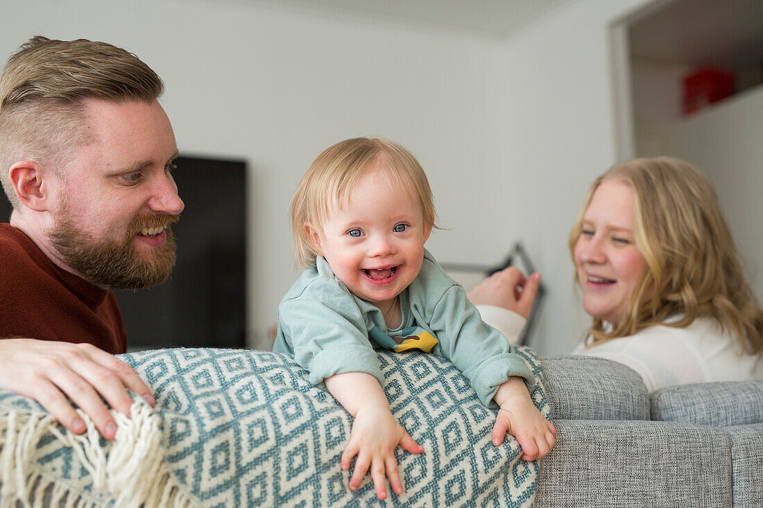 Eltern spielen mit lächelndem Baby Eltern mit Baby mit Down-Syndrom lesen ein Buch auf Sofa auf Sofa im Wohnzimmer