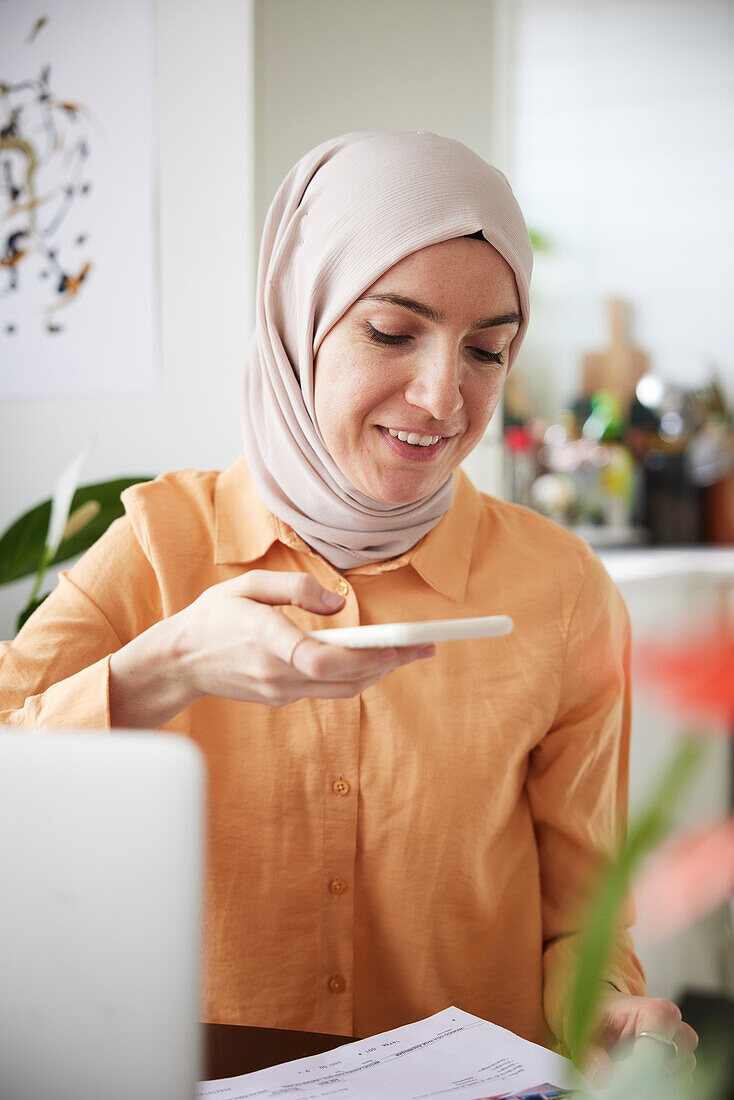 Smiling woman with hijab using cell phone and scanning bills