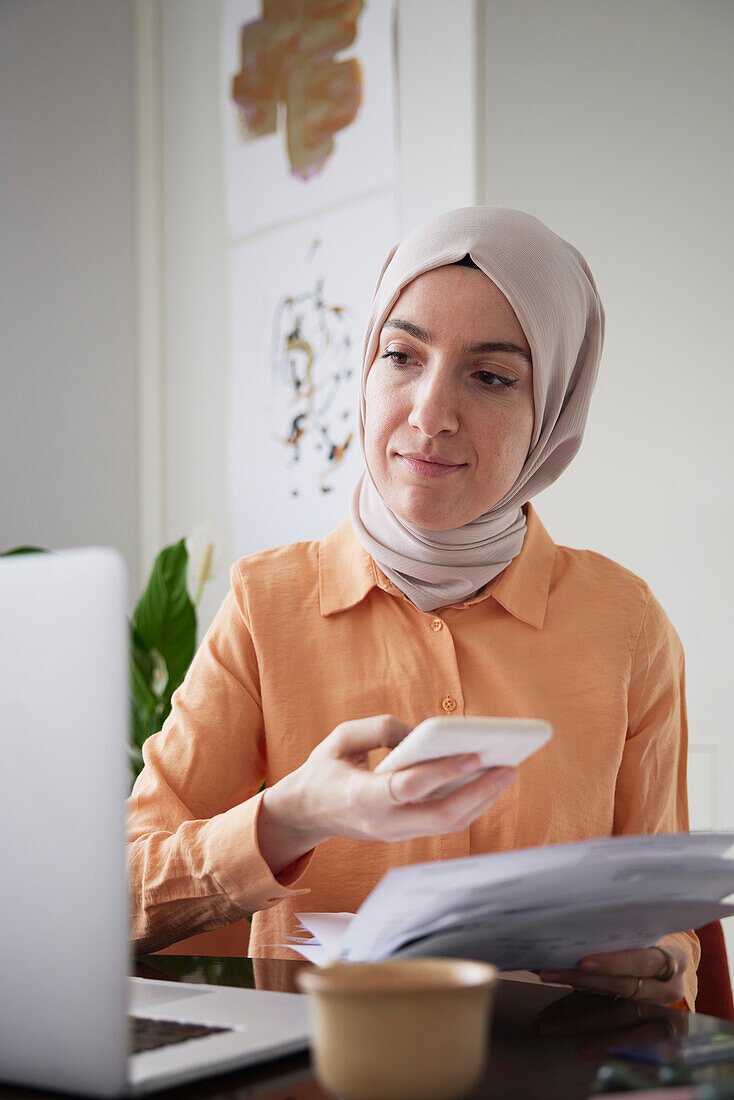 Smiling woman with hijab using cell phone and scanning bills