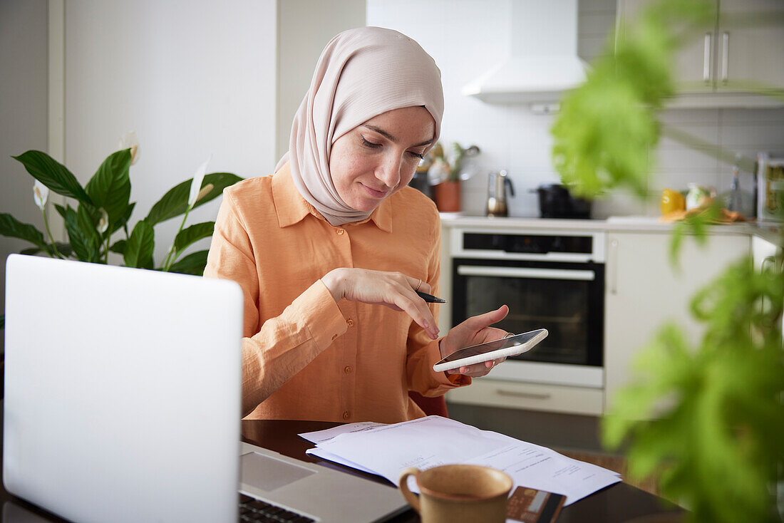 Lächelnde Frau mit Hijab telefoniert während der Heimarbeit in der Küche