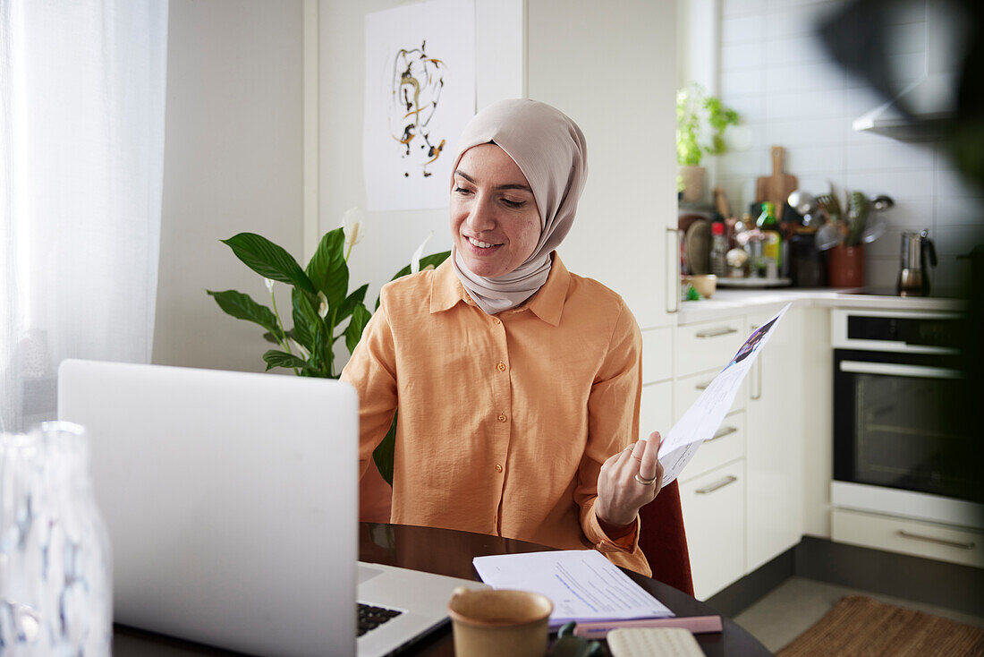 Lächelnde Frau mit Hidschab prüft Rechnungen zu Hause