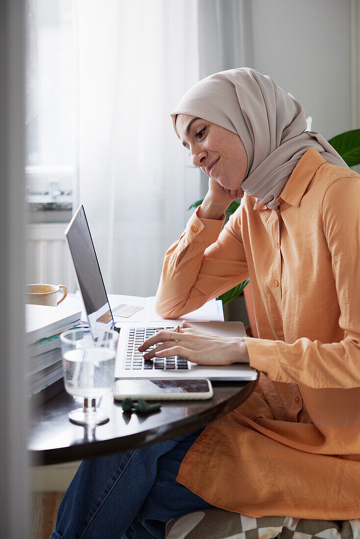 Lächelnde Frau mit Hidschab bei der Heimarbeit am Laptop