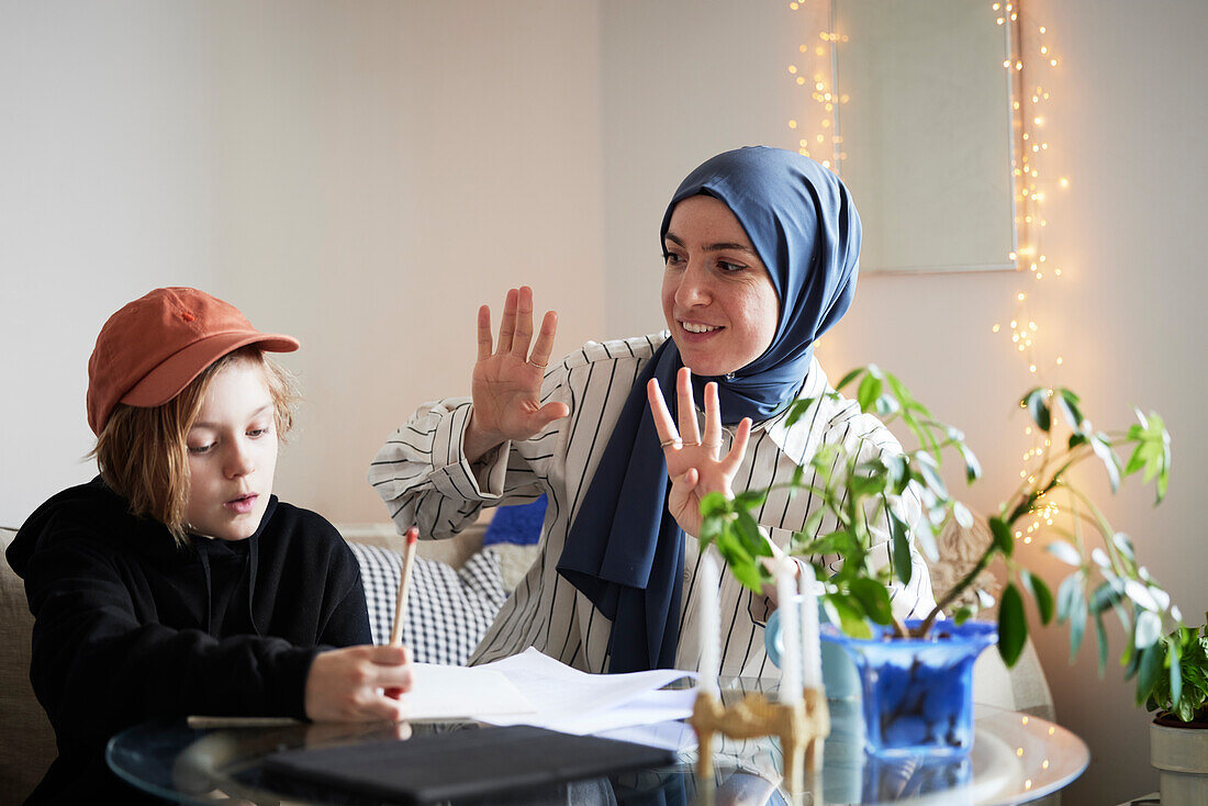Mother wearing hijab holds up fingers while helping son doing math homework