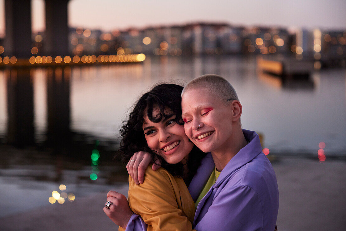 Happy young female friends or gay couple hugging, river in background