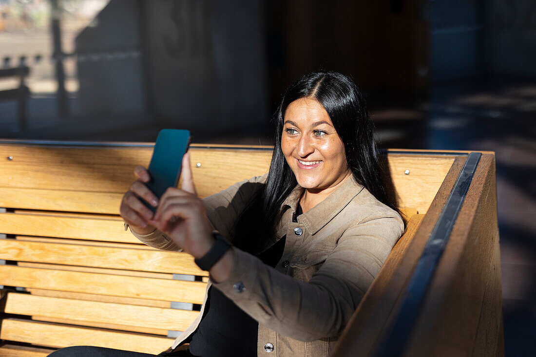 Lächelnde Frau sitzt auf dem Rücken im Bahnhof und macht ein Selfie