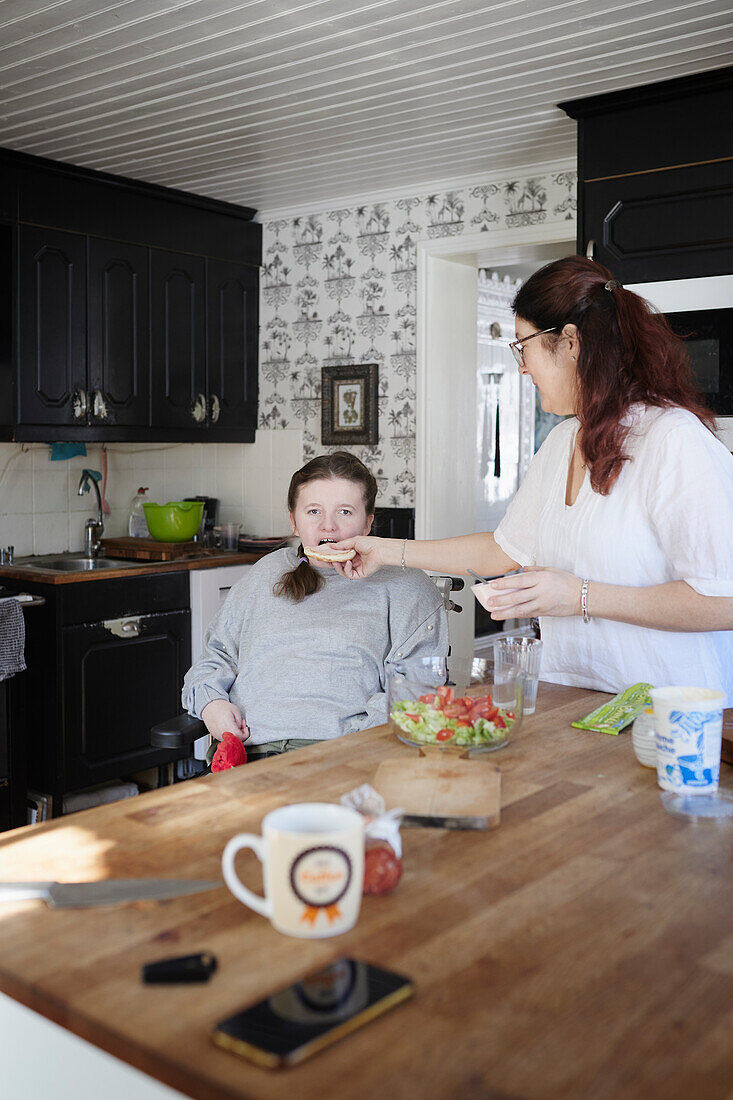 Mutter füttert behinderte jugendliche Tochter im Rollstuhl am Küchentisch