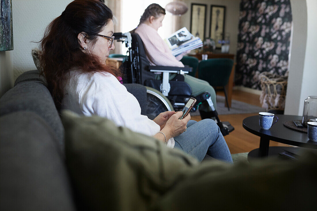 Woman using cell phone on sofa, disabled teenage daughter in wheelchair in background