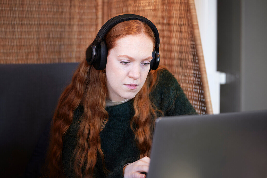 Junge Frau mit Kopfhörern, die Musik oder einen Podcast hört, während sie mit dem Laptop lernt