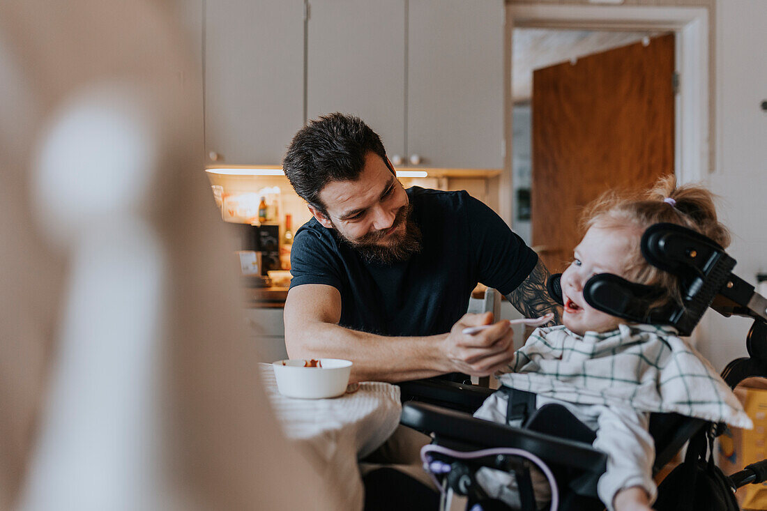 Smiling father feeding disabled child in wheelchair in kitchen