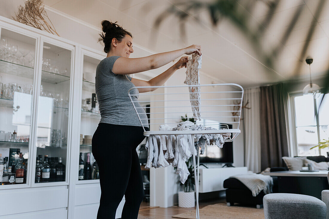 Schwangere Frau bei der Hausarbeit und beim Aufhängen von Babykleidung auf dem Wäscheständer