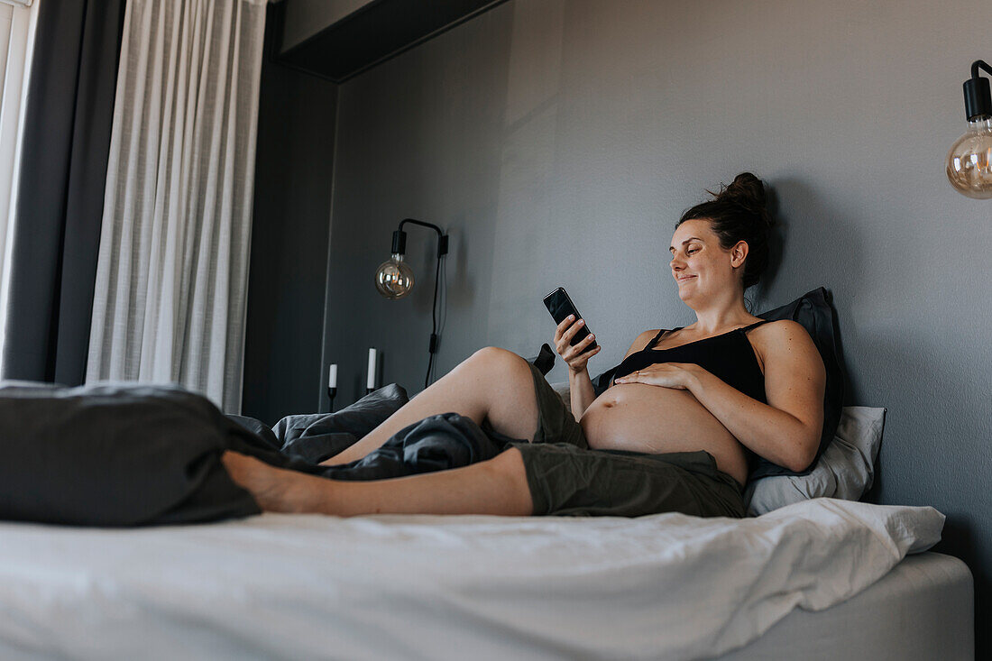 Glückliche schwangere Frau entspannt sich im Bett und benutzt ihr Telefon