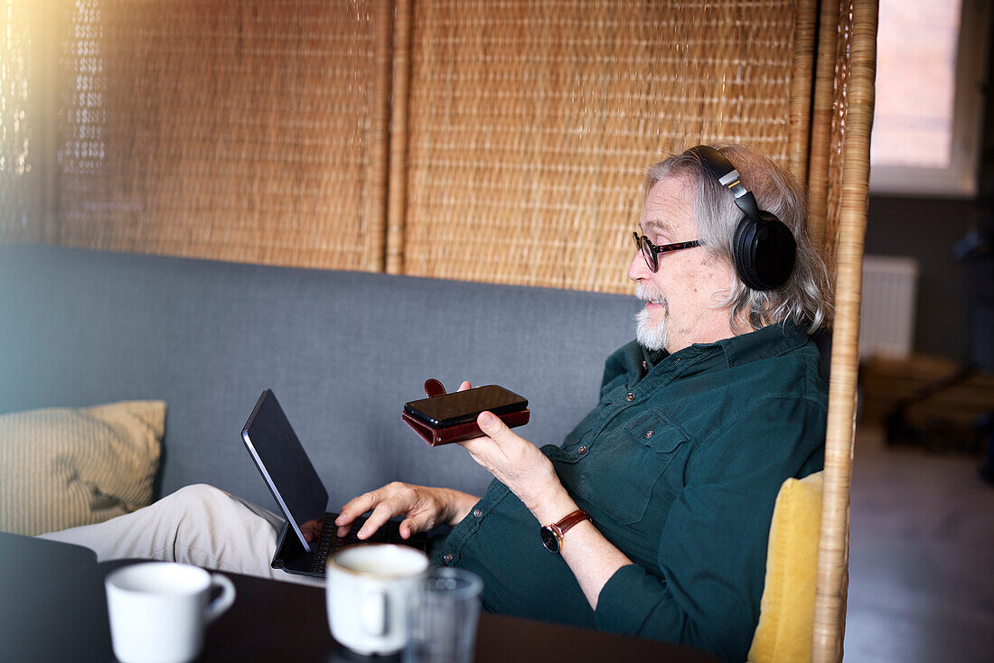 Älterer Mann mit Kopfhörern, Telefon und Tablet in einem Café
