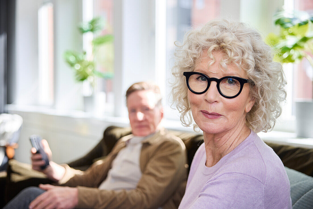 Lächelnde reife Frau schaut in die Kamera, Mann sitzt im Hintergrund