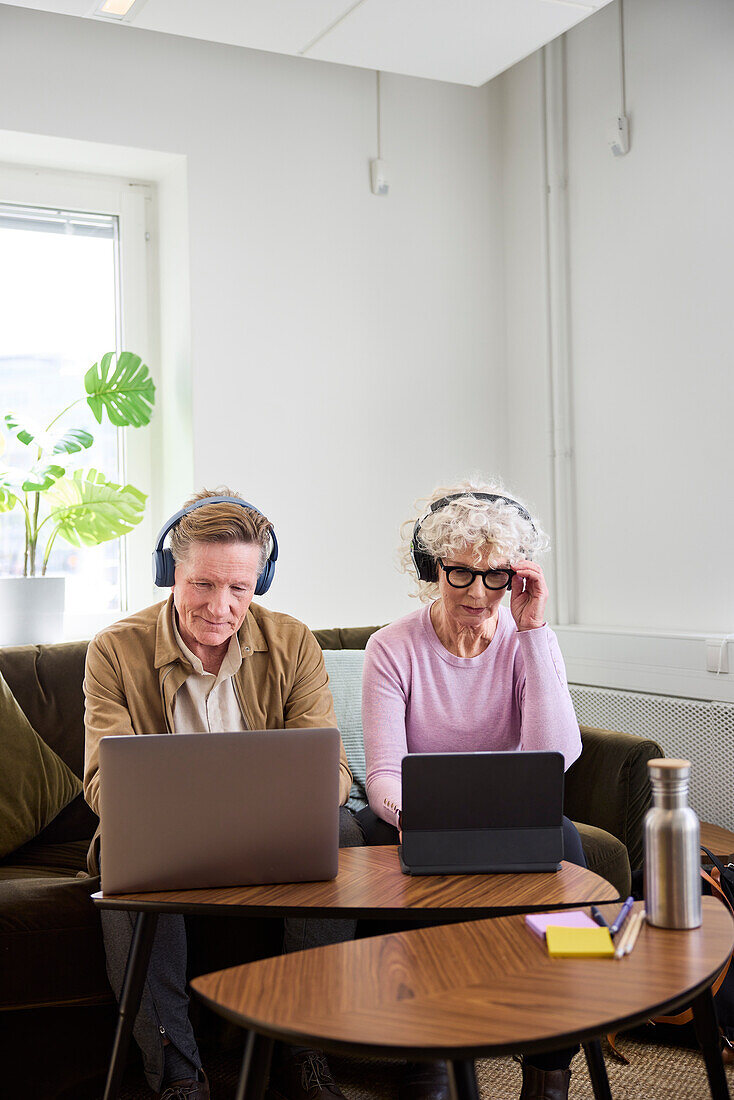 Älterer Mann und Frau sitzen im Wohnzimmer und benutzen Laptop und digitales Tablet, um Podcast zu bearbeiten