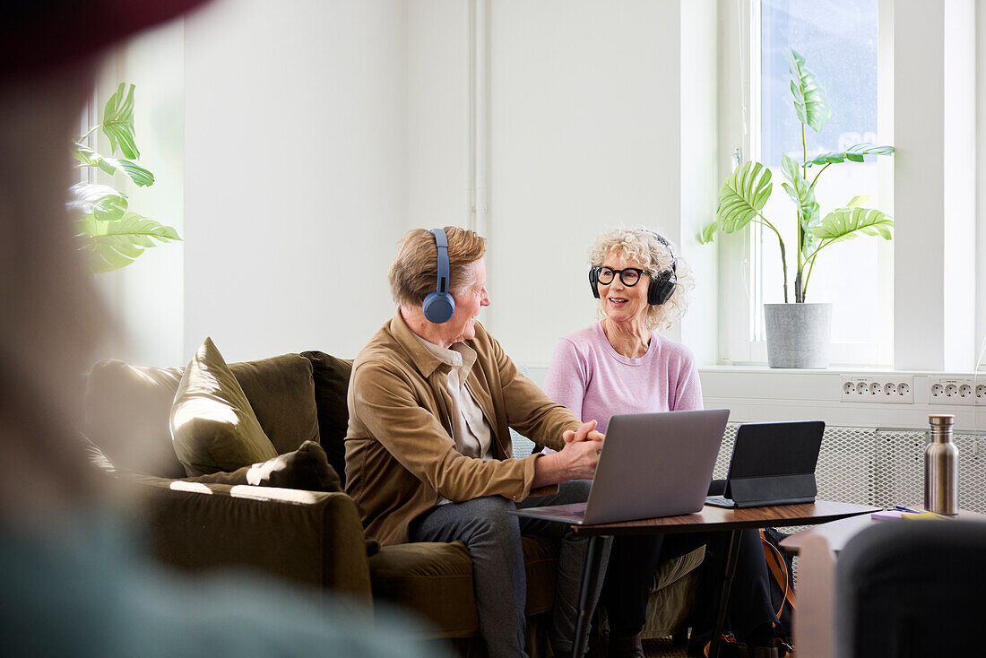 Älterer Mann und Frau im Gespräch, während sie im Wohnzimmer sitzen und mit Laptop und digitalem Tablet einen Podcast bearbeiten