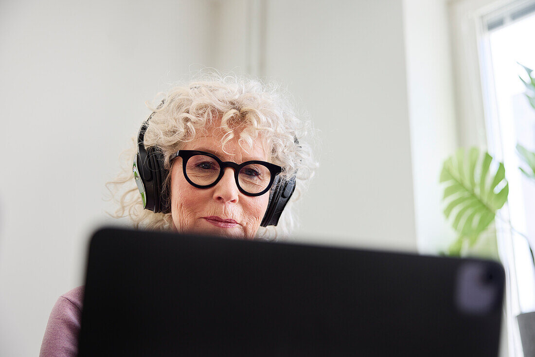 Lächelnde ältere Frau sitzt mit Kopfhörern auf und benutzt einen Laptop