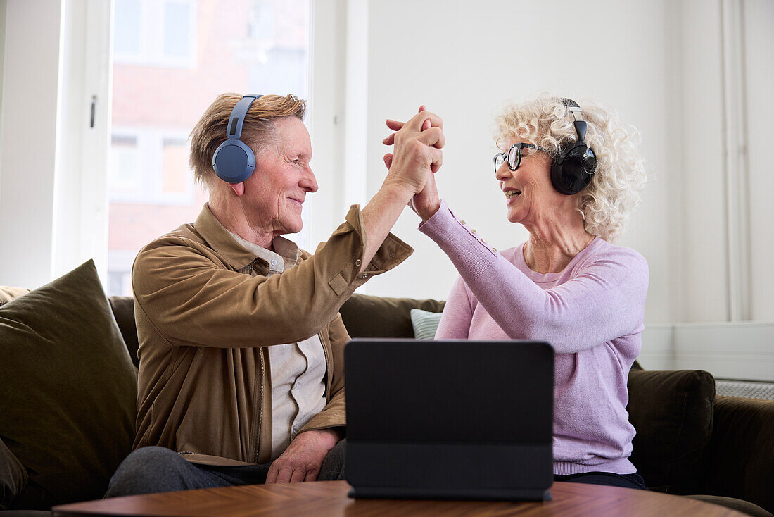 Älterer Mann und Frau geben sich gegenseitig ein High Five, während sie im Wohnzimmer vor einem digitalen Tablet sitzen