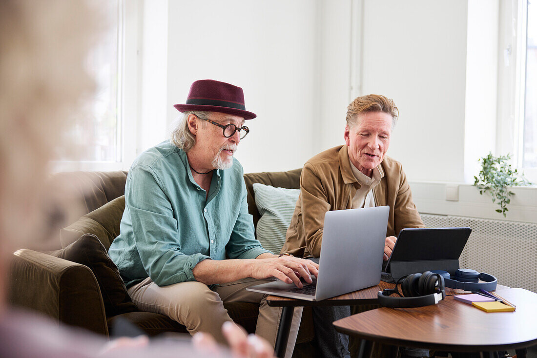 Ältere Männer reden miteinander, während sie im Wohnzimmer sitzen und ein digitales Tablet und einen Laptop benutzen, um einen Podcast zu bearbeiten