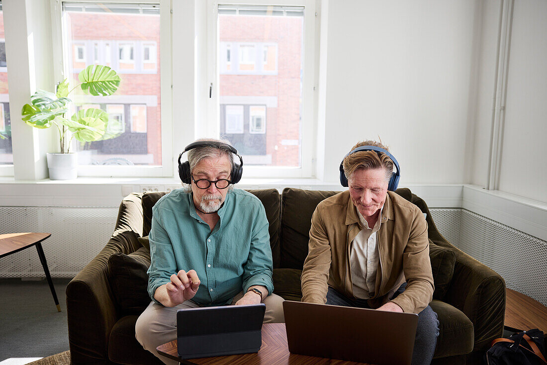 Ältere Männer sitzen im Wohnzimmer und benutzen ein digitales Tablet und einen Laptop, um einen Podcast zu bearbeiten