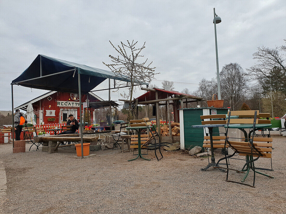 A roadside stall with tables and chairs in a rural setting, a man seated on a bench, a pile of logs.