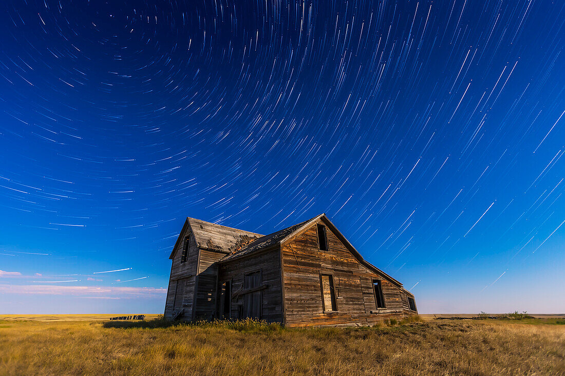 Zirkumpolare Sternenschweife kreisen über einem alten rustikalen und verlassenen Haus in der Nähe von Bow Island, Alberta, mit Beleuchtung durch den fast vollen Mond. Kassiopeia ist nahe der Mitte. Polaris ist oben links.