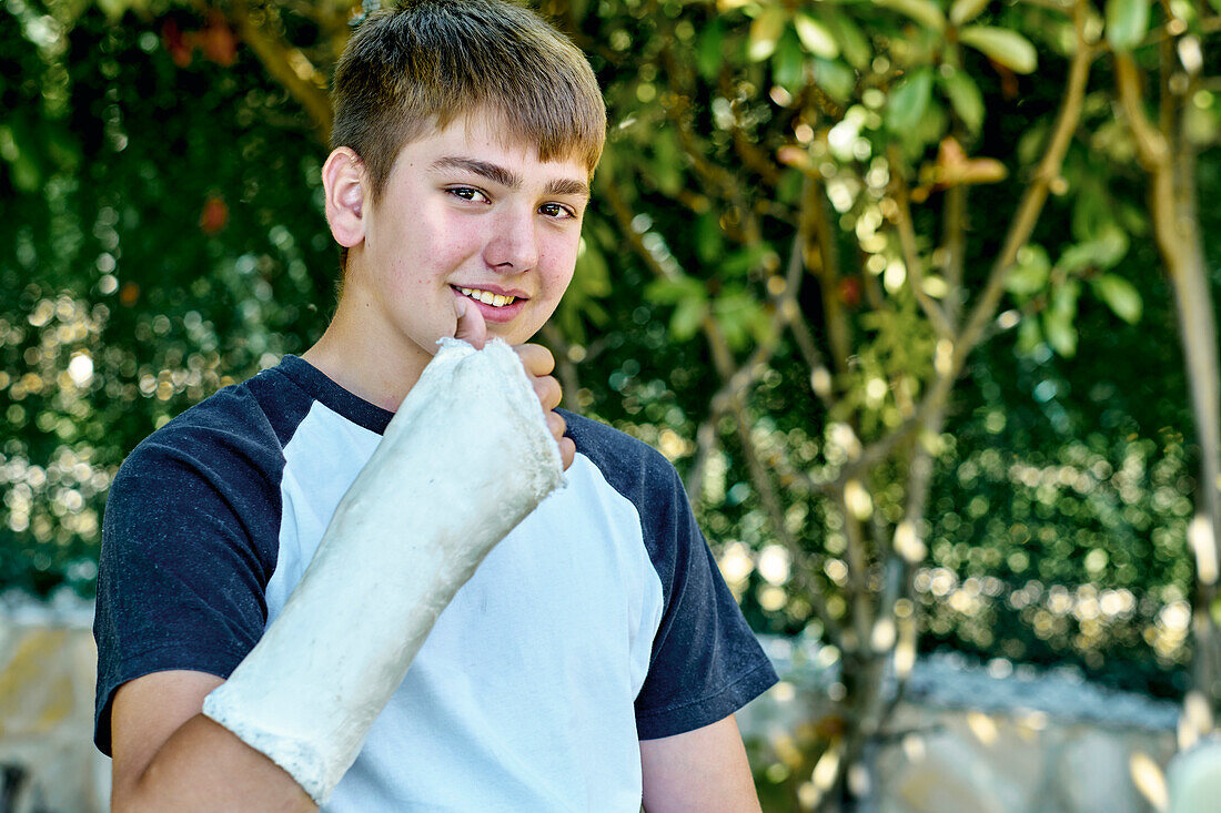 Porträt eines jungen kaukasischen Jungen mit gebrochenem und eingegipstem Arm, der auf einem Stuhl im Freien in einem Garten sitzt. Lebensstil-Konzept.