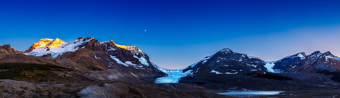 The last rays of the setting Sun catch the peaks around the Columbia Icefields in Jasper National Park, Alberta, on July 27, 2020. The waxing quarter Moon shines over Mount Andromeda and sunlight illuminates the glacier on Mount Athabasca at left. The famous Athabasca Glacier itself is at centre. Snow Dome Glacier is at right. The meltwater lake in the middle distance is Sunwapta Lake.