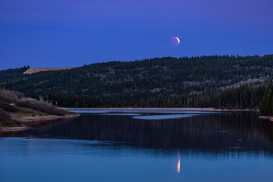 Der verfinsterte Vollmond geht am 15. Mai 2022 über dem Reesor Lake im Cypress Hills Interprovincial Park, Alberta, auf. Dies war in der letzten Phase der partiellen Verfinsterung, wobei ein Teil der Mondscheibe noch vom direkten Sonnenlicht beleuchtet wurde, während der Rest im roten Kernschatten lag.