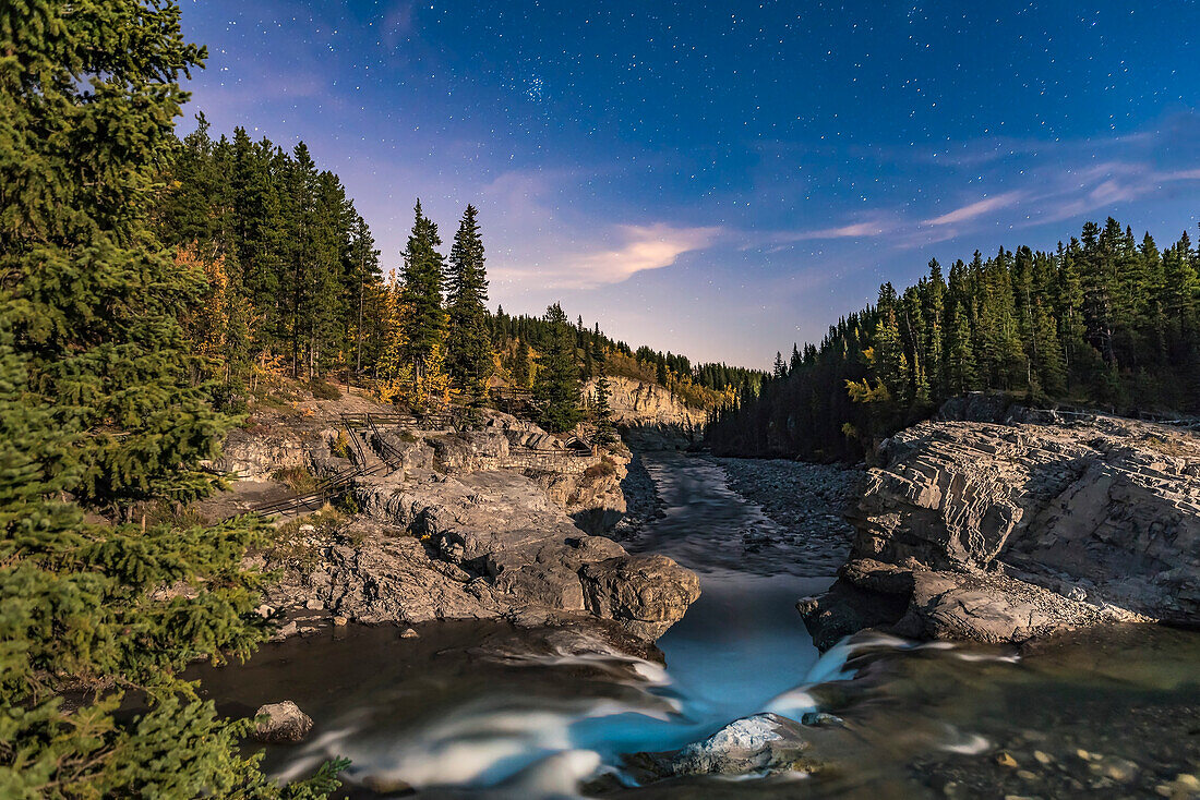 Der Pleaides-Sternhaufen (die Sieben Schwestern) und die Sterne des Spätherbstes und Winters, die über den Elbow Falls im Kananaskis Country in Alberta in einer mondhellen Nacht aufgehen, wobei der zunehmende Gibbous-Mond für die Beleuchtung sorgt.