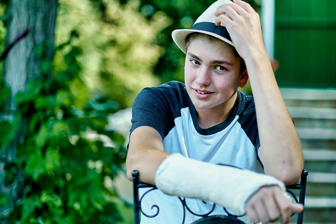 Portrait of young caucasian boy with a broken and cast arm wearing a hat and sitting in a chair outdoor in a garden. Lifestyle concept.