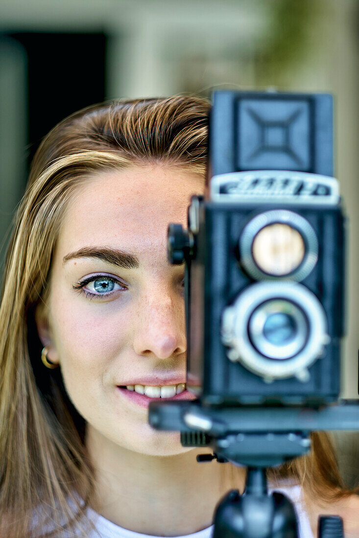 Porträt einer jungen schönen kaukasischen Frau in den 20ern mit blauen Augen, die mit einer alten Vintage-Kamera auf einem Stativ im Freien fotografiert. Lebensstil-Konzept.