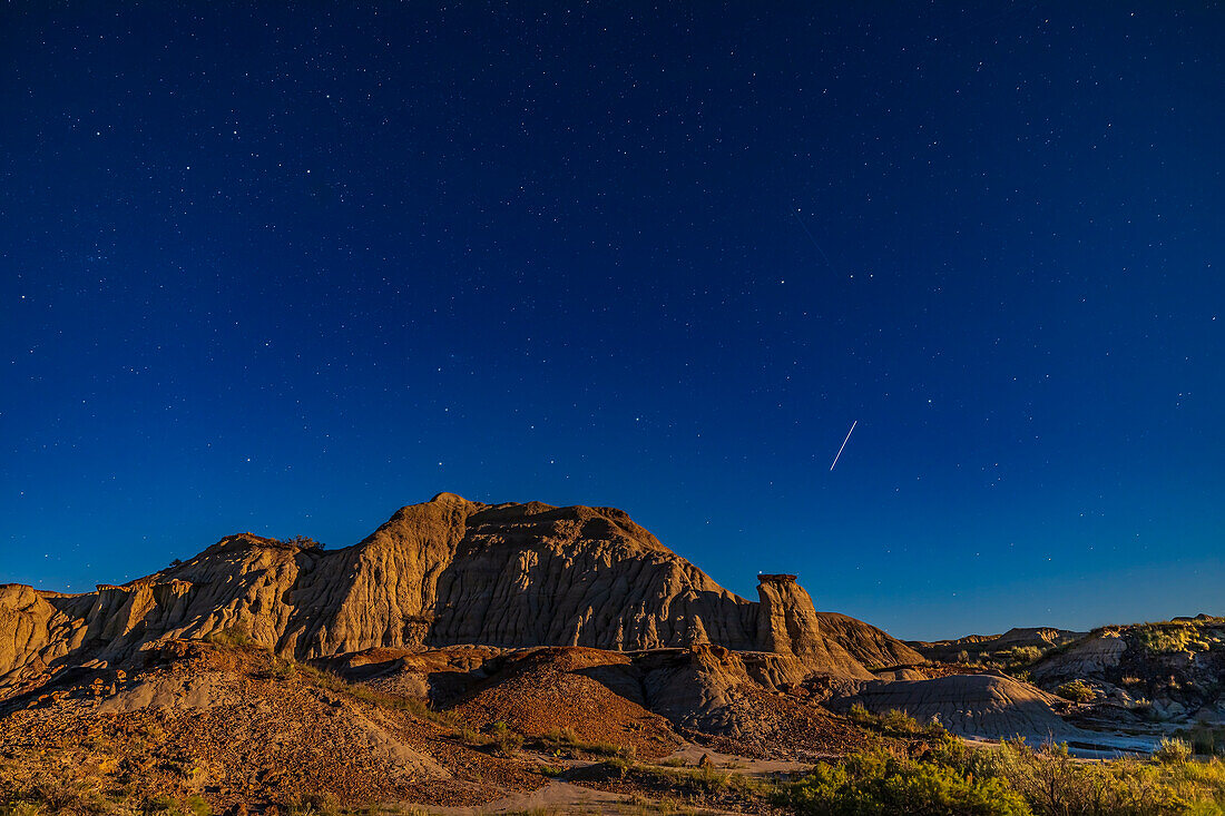 Die Internationale Raumstation (ISS) fliegt am 12. Juli 2022, kurz vor Mitternacht, in östlicher Richtung über die mondbeschienenen Badlands im Dinosaur Provincial Park, Alberta, hinweg. Das Bild umrahmt die Sterne Kassiopeia (oben links), Perseus (links), Andromeda (Mitte) und Pegasus (rechts). Einige andere schwächere Trabanten sind ebenfalls auf dem Bild zu sehen. Das Licht des fast vollen Mondes lässt den Himmel blau und den Vordergrund in warmen Farben erstrahlen.