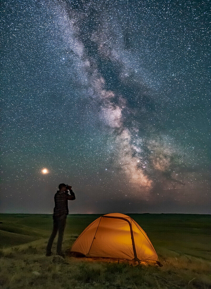 Ein Parkdolmetscher posiert für eine Szene im Grasslands National Park, Saskatchewan, bei der Sternbeobachtung mit dem Fernglas unter der Milchstraße in einer dunklen mondlosen Nacht. Grasslands eignet sich perfekt für die Sternenbeobachtung, da es ein Dark Sky Preserve ist und der Horizont weit und unverbaut ist.