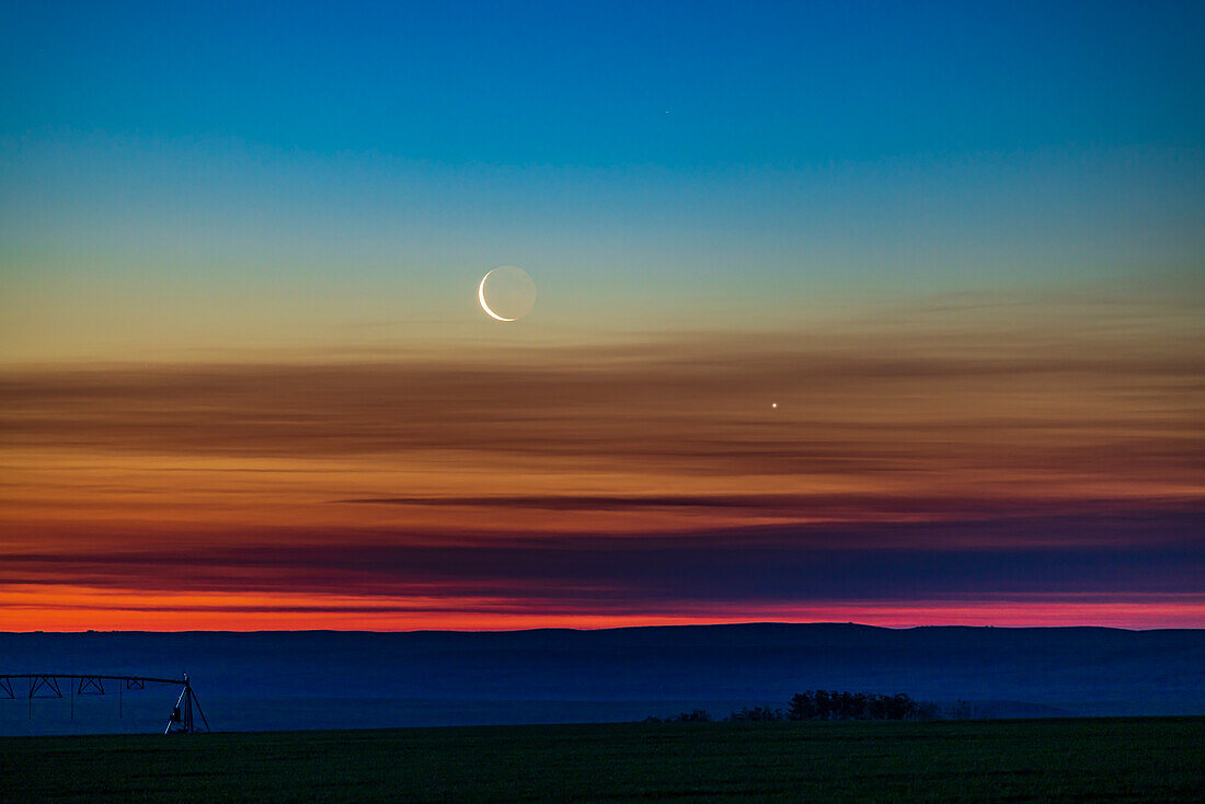 Die Konjunktion der abnehmenden Mondsichel mit der Venus, als sie am 26. Juni 2022 tief am nordöstlichen Morgenhimmel aufsteigen, aufgenommen von zu Hause in Süd-Alberta, 51° nördlicher Breite. Der Himmel zeigt einen wunderbaren Farbübergang vom Orange am Horizont über das Spektrum bis hin zu den Blautönen am oberen Rand.