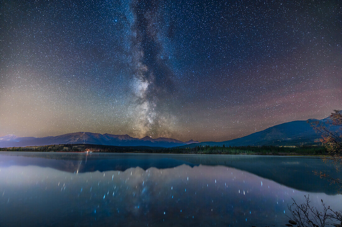 Die sommerliche Milchstraße, die sich in einer Nacht Mitte Oktober im relativ ruhigen Wasser des Pyramid Lake im Jasper National Park spiegelt. Die Jasper Sky Tram fügt die Lichter auf dem Whistler Peak hinzu. Der Himmel ist durch das Glühen der Luft rot gefärbt. Die Lichter der Stadt Jasper, die 2022 immer noch größtenteils aus nicht abgeschirmten Natriumdampflampen bestehen, fügen das Himmelsleuchten links hinzu. Altair ist der helle Stern ganz oben. Der rote Lagunennebel geht gerade hinter der Gebirgskette unter. Der leichte Wind hat das Wasser so gekräuselt, dass eine perfekte Reflexion ni