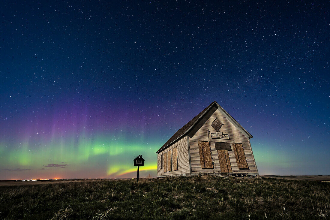 Das Liberty Schoolhouse von 1910, eine klassische Einraumschule der Pioniere, in der Prärie von Alberta unter dem Sternenhimmel einer Frühlingsnacht, während im Norden ein Polarlicht tanzt. Das Mondlicht sorgt für die Beleuchtung. Kassiopeia befindet sich über der Schule. Polaris ist ganz oben.