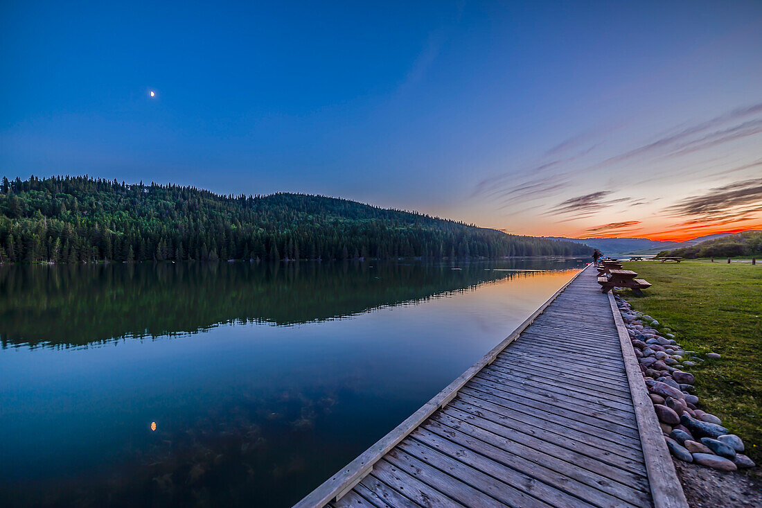 Der Viertelmond spiegelt sich im Wasser des Reesor Lake, Alberta im Cypress Hills Interprovincial Park. Aufgenommen am 5. Juli 2014. Dies ist mit dem 14mm Rokinon Objektiv und der Canon 6D bei ISO800 aufgenommen. Es handelt sich um einen Stapel mit hohem Dynamikbereich aus 6 Aufnahmen von 1/15 bis 0,6 Sekunden, die kurz vor der Verwendung der Kamera für einen bewegungsgesteuerten Zeitraffer aufgenommen wurden. Der Mond stand in Konjunktion mit Mars (rechts vom Mond) und Spica (links vom Mond), aber in der hellen Dämmerung sind sie hier nicht zu sehen.