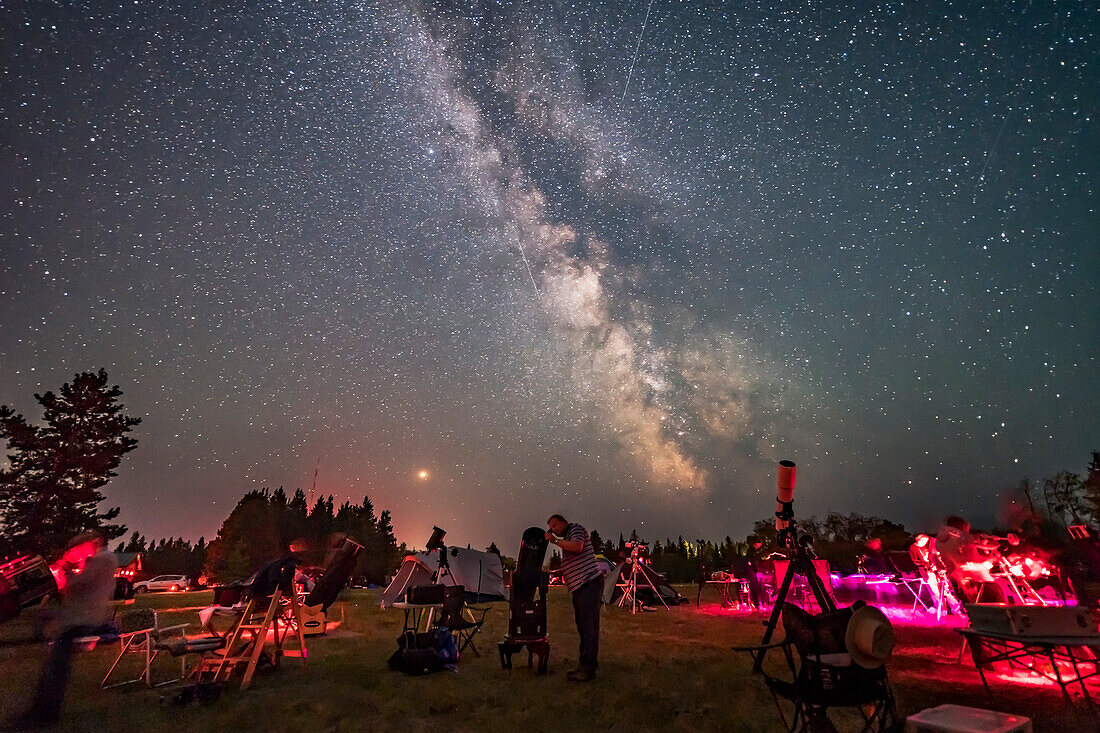 Ein Beobachter blickt mit seinem Dobson-Spiegelteleskop bei der Saskatchewan Summer Star Party am 9. August 2018 in den Cypress Hills im Südwesten von Saskatchewan, im Cypress Hills Interprovincial Park, einem Dark Sky Preserve, zum Himmel. Die Milchstraße leuchtet im Süden. Der Rauch am Himmel verdeckt den Horizont etwas.