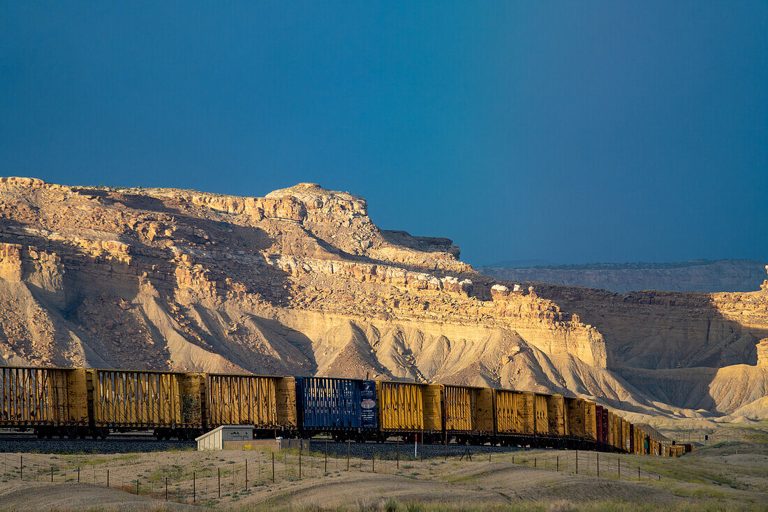 A line of centerbeam rail cars in the Green River Desert of Utah with spotlighting on the Book Cliffs behind.