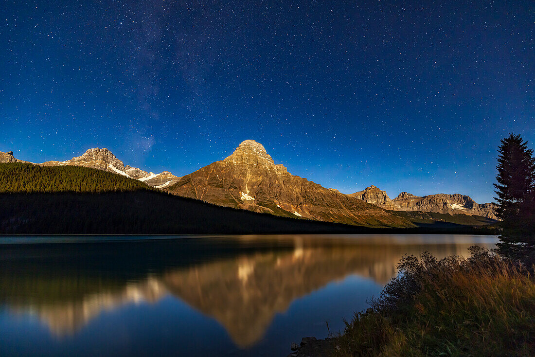 Der Mount Cephren am Lower Waterfowl Lake, im Licht des abnehmenden Mondes, der die Gipfel, aber nicht den Vordergrund beleuchtet. Dies ist eine Aufnahme vom Aussichtspunkt am Seeufer des Icefields Parkway im Banff National Park, Alberta. Ich habe es in einer sehr klaren Nacht am 13. Oktober 2022 aufgenommen. Der Wind reichte aus, um das Wasser zu kräuseln und Sternenreflexe zu verwischen. Die Milchstraße befindet sich links von Cephren, geht aber im heller werdenden Mondhimmel unter.