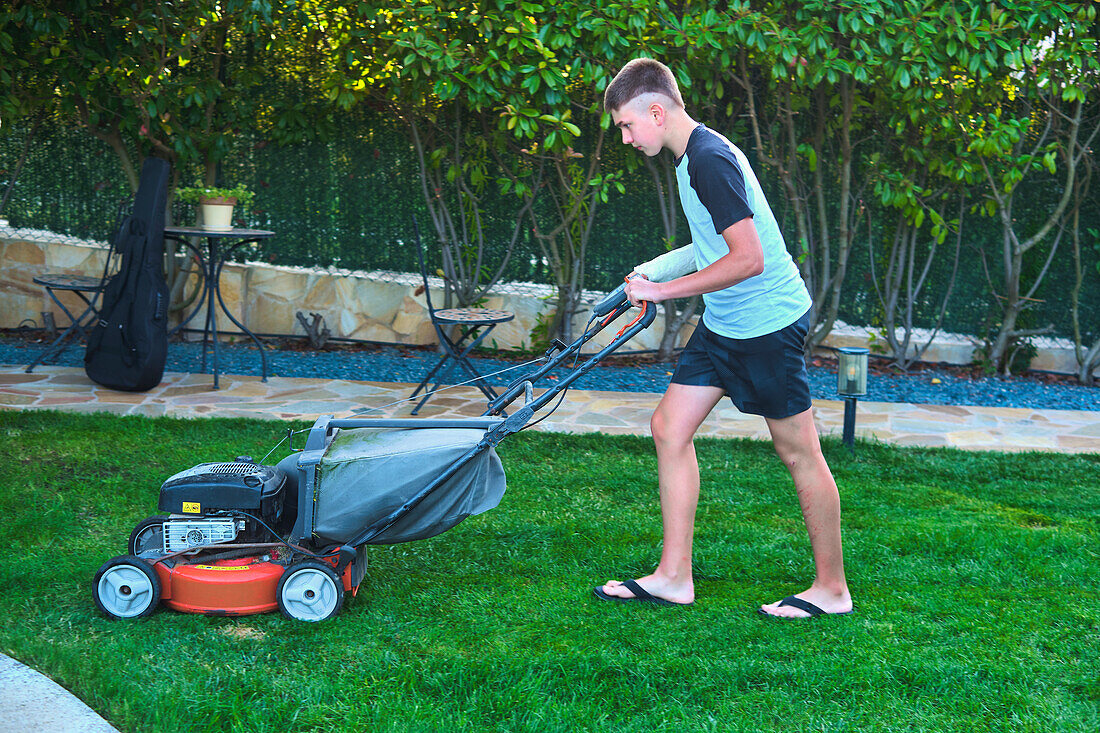 Portrait of a young caucasian boy passing the mower through in the garden in a country house. Lifestyle concept.
