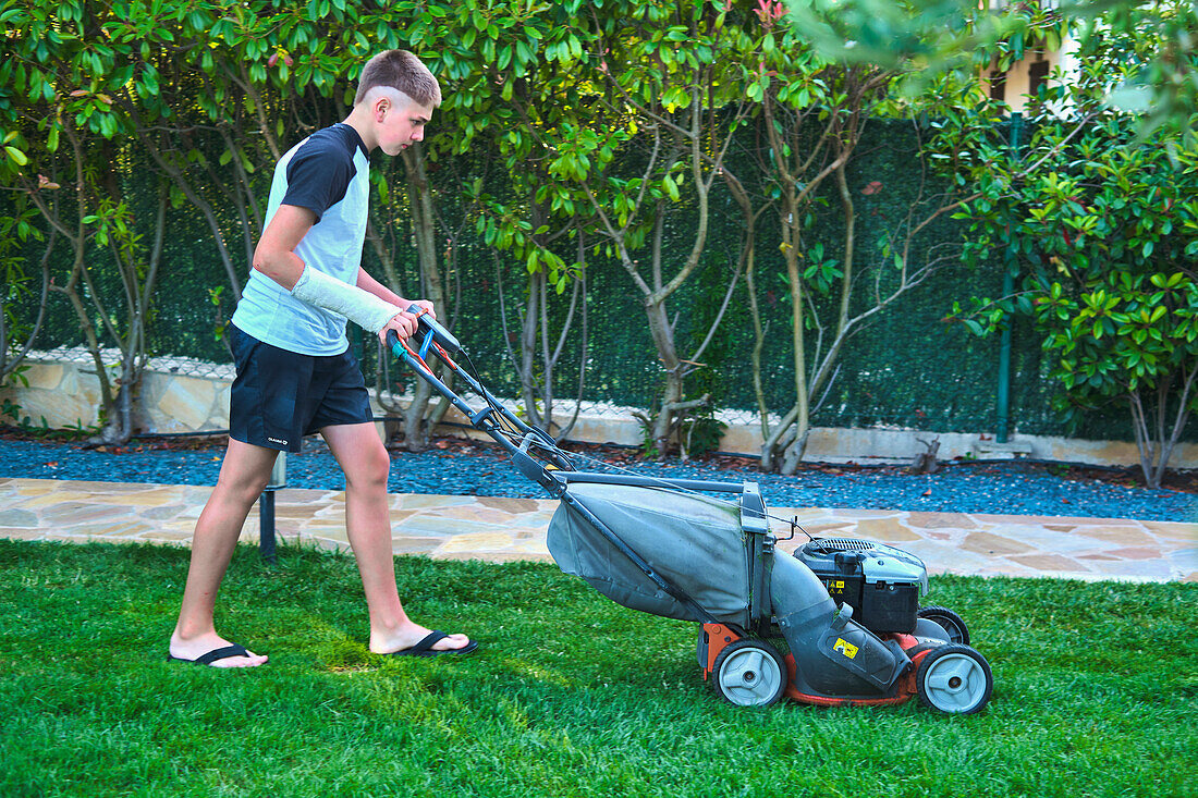 Portrait of a young caucasian boy passing the mower through in the garden in a country house. Lifestyle concept.