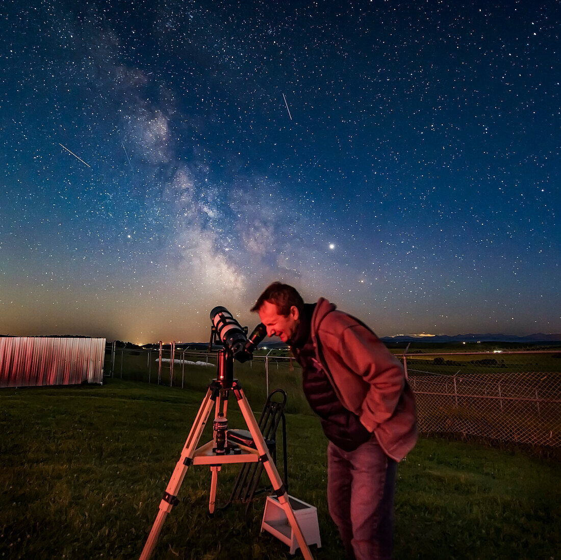 Mark gazing at a target, M22, in the Milky Way with his TeleVue 127 refractor at the annual Rothney Observatory Milky Way Nights for July 25, 2019. Several satellite trails mark the sky. Jupiter (brightest at right) and Saturn (at left) flank the Milky Way.