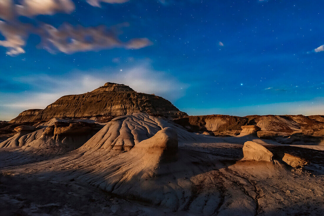 The eroding formations of Dinosaur Provincial Park, Alberta, lit by the rising gibbous Moon, off camera at left, on April 21/22, 2019. This is looking west, with the stars of the winter sky setting. Procyon is at right. Aphard in Hydra is above the hill.