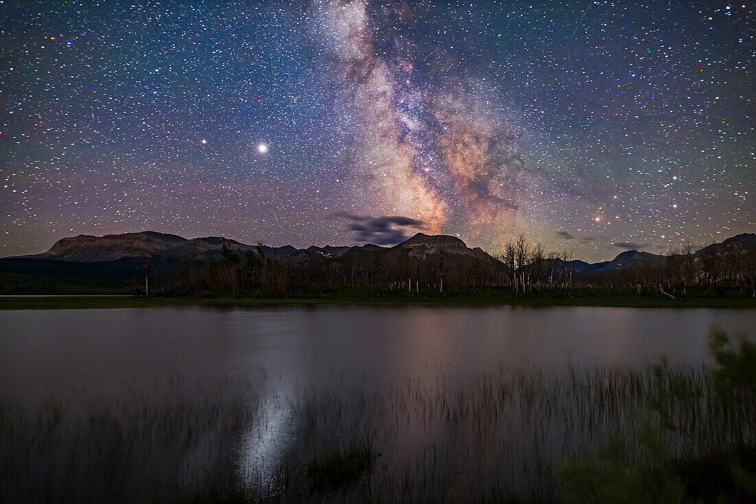 The galactic core area of the Milky Way over Maskinonge Pond in Waterton Lakes National Park, Alberta. Jupiter is the bright object at left, with Saturn dimmer to the left (east) of Jupiter. In the summer of 2020 the two planets were close together in the summer sky. Jupiter provides the glitter path on the water. Antares and Scorpius are to the right. Sagittarius is at centre. This was July 13-14, 2020.