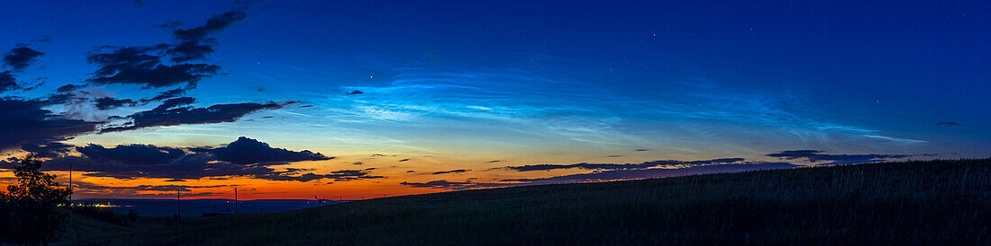 A panorama of NLCs – noctilucent clouds – aka PMCs or polar mesospheric clouds, taken on June 20, 2022 about 11:40 pm MDT, from home in southern Alberta. After many cloudy nights and some clear nights with no NLCs, this was my first sighting since June 1. The bright luminous mesospheric clouds lit by sunlight at high altitude contrast with the dark tropospheric clouds to the left at low altitude.