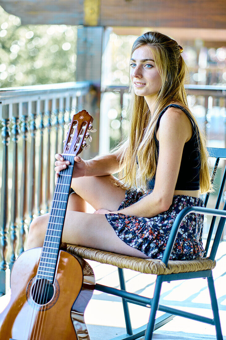 Portrait of a young beautiful caucasian woman in her 20´s with long hair and blue eyes with a guitar on the porch of a country house. Lifestyle concept.
