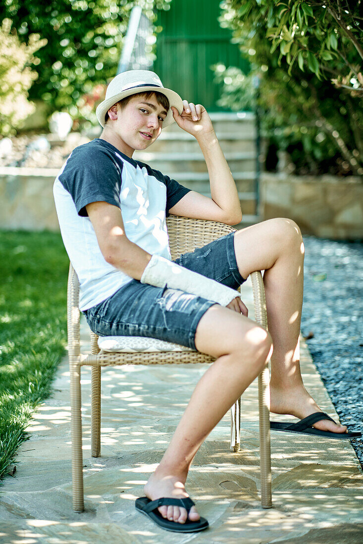 Porträt eines jungen kaukasischen Jungen mit gebrochenem und eingegipstem Arm, der einen Hut trägt und in einem Stuhl im Freien in einem Garten sitzt. Lebensstil-Konzept.